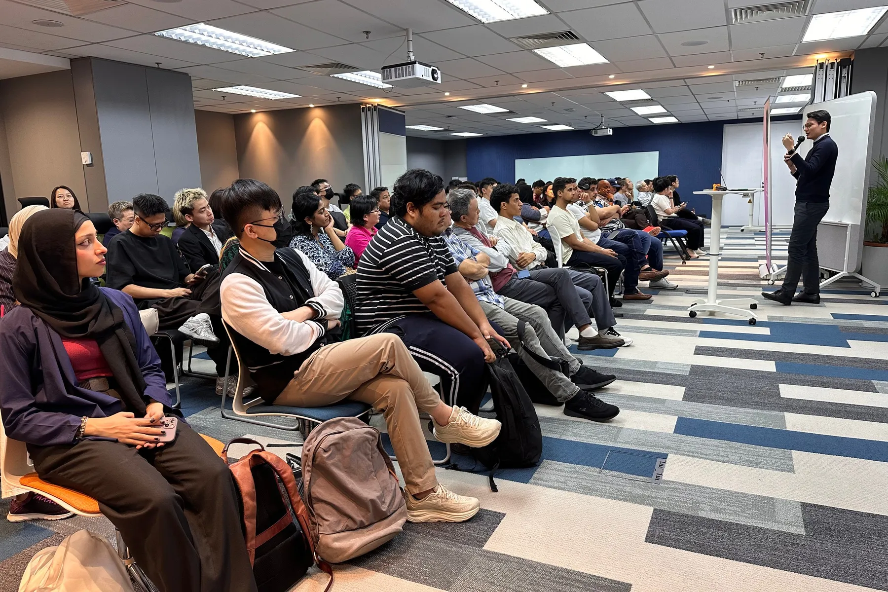 A speaker with a microphone talks to rows of seated attendees in a meeting room