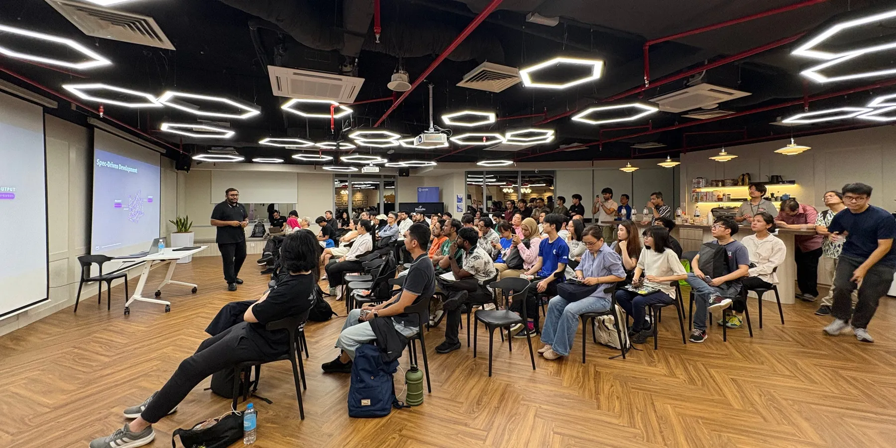 A speaker presents to a full room of attendees under hexagon-shaped ceiling lights